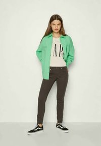 Camisa verde de botões sobre uma T-shirt branca com a inscrição "GAP", combinada com umas calças skinny cinza-escuras e sapatilhas pretas.