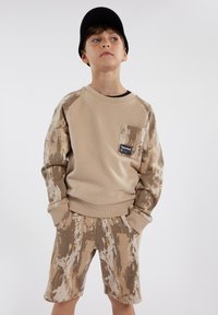Sweatshirt beige avec des manches et une poche à motifs, associé à un short beige arborant un design abstrait similaire. Tissu doux, coupe décontractée.