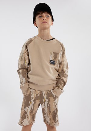 Sweatshirt beige avec des manches et une poche à motifs, associé à un short beige arborant un design abstrait similaire. Tissu doux, coupe décontractée.