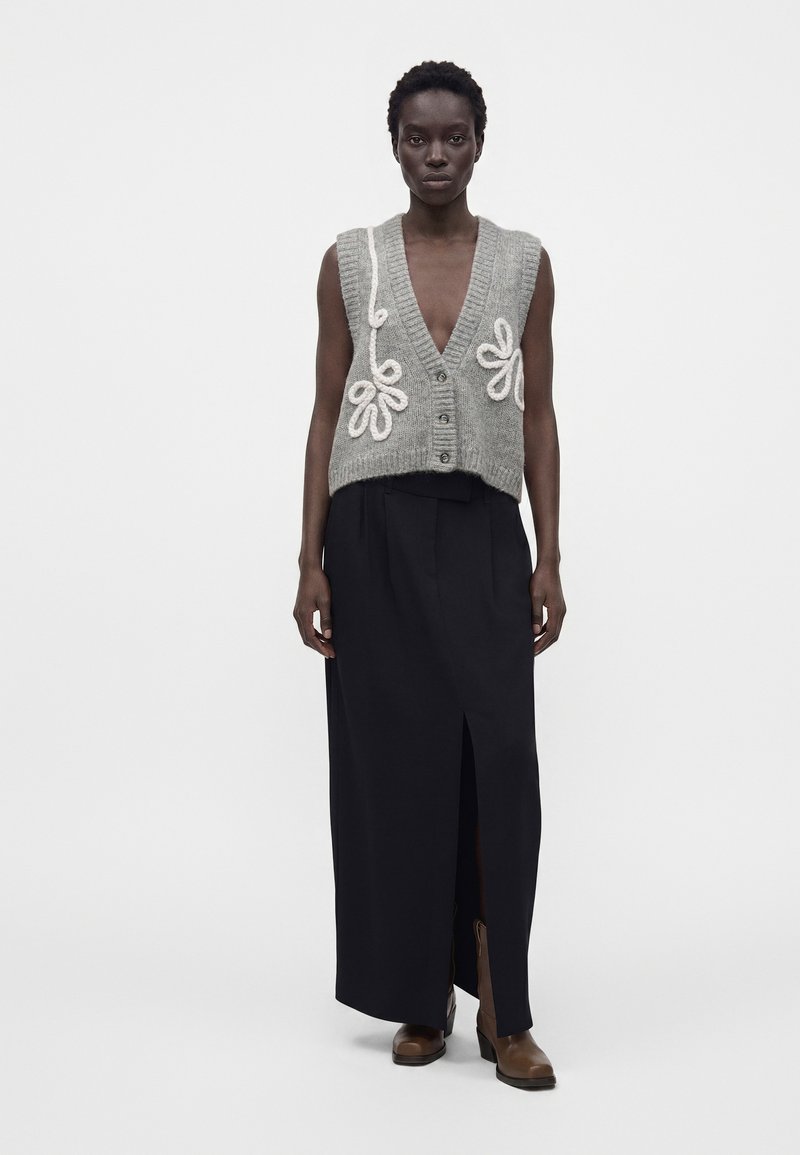 Femme portant un gilet gris en maille boutonné avec des détails floraux blancs, une longue jupe noire fendue à l'avant, et des bottines marron, debout immobile.