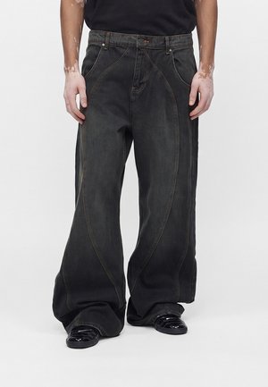 Άνδρας που φοράει μαύρα τζιν σε μεγάλο μέγεθος με καμπυλωτές ραφές και μαύρα γυαλιστερά παπούτσια, στέκεται σε λευκό φόντο.