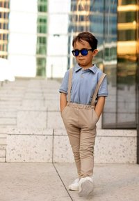 Jasnoniebieska koszulka polo z krótkimi rękawami, beżowe spodnie z szelkami, białe trampki i niebieskie okulary przeciwsłoneczne. Stoi na teksturowanej powierzchni.