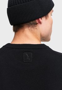 Muž s čiernou pletenou čiapkou a čiernym sveterom so malou vyšitou obdĺžnikovou nášivkou na hornom chrbte.