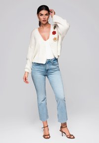 Vit stickad cardigan med blommönster, bärs över en åtsittande vit topp och ljusblå avskuren jeans, kombinerad med bruna remsandaler.