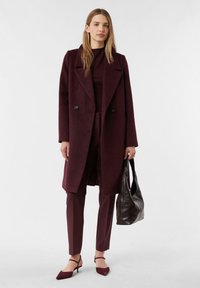 Kobieta ubrana w bordowy płaszcz, pasujące spodnie i szpilki, trzyma ciemno-brązową skórzaną torbę na tle jasnego, jednolitego tła.