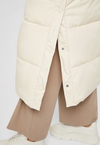 Kræmfarvet pufferjakke med sideslids, der har trykknapper og et elegant finish; parret med beige bredbenede bukser og hvide sneakers.