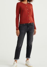 Bränd orange cardigan med rund hals och knäppning, tillsammans med mörka denimjeans, uppvikta ärmslut, och vita sneakers.