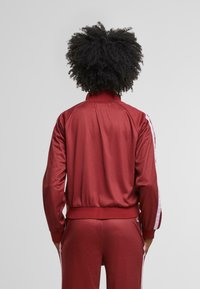 Červená sportovní bunda s texturovaným povrchem, vysokým límcem a bílými pruhy po stranách. Design zahrnuje vypasovaný pas a dlouhé rukávy.