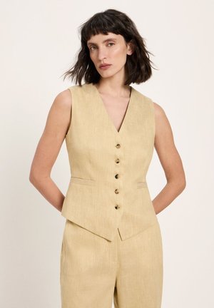 Frau mit schulterlangem dunklem Haar, die eine ärmellose, beige, geknöpfte Weste und passende Hose vor einem schlichten Hintergrund trägt.