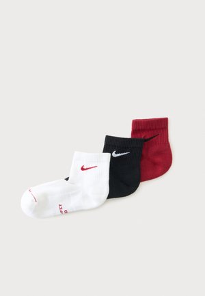 Trois paires de chaussettes chevilles Nike en blanc avec logo rouge, noir avec logo blanc et rouge avec logo noir, disposées en chevauchement sur un fond uni.
