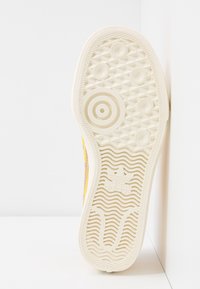 Gummisulor i krämfärg med texturerat greppmönster, cirkulära detaljer och varumärkeslogotyp. Sidan visar en ljusgul ovandel.