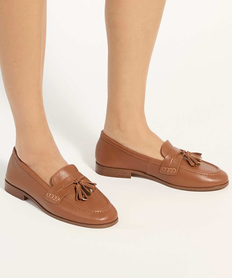 Jambes portant des mocassins en cuir marron à pampilles avec des talons bas empilés sur un fond clair.