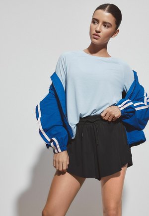 Jeune femme posant vêtue d'une chemise bleu clair, d'une veste zippée bleue avec des rayures blanches, et d'un short noir sur un fond uni.