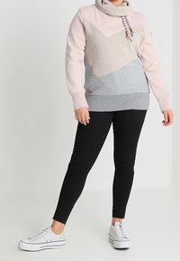 Sudadera ligera en rosa, gris y jaspeado con patrones geométricos; cuello alto, cordón y detalle de logo en el dobladillo. Jeans pitillo negros y zapatillas.
