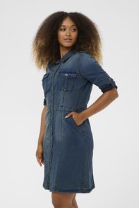 Kvinna med lockigt hår klädd i en knappad denimklänning med uppvikta ärmar, stående med en hand i en sidoficka och tittande åt sidan.