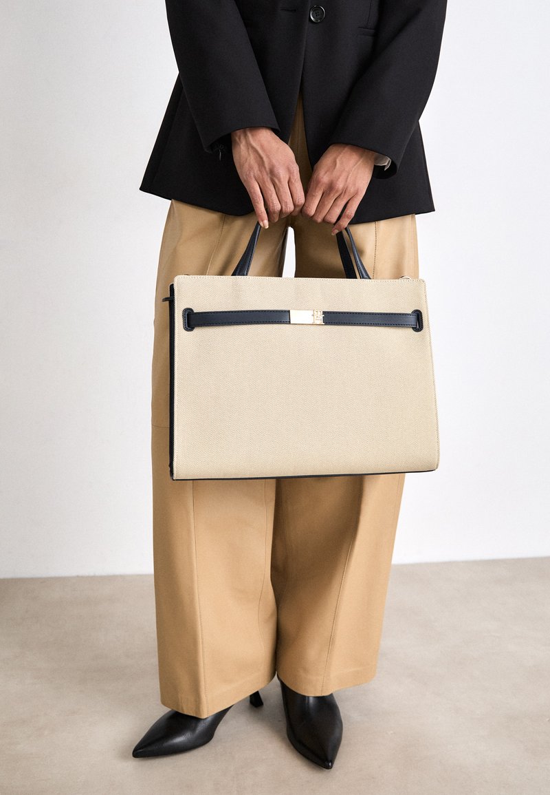 Beige gestructureerde totebag met textuurafwerking, donkerblauwe handvatten, gouden hardware-accent, vastgehouden door een persoon in een zwart colbert en beige broek.