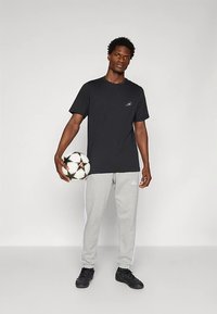 Μαύρο μπλουζάκι και γκρι φόρμα με ρίγες στο πλάι, κρατώντας μία μπάλα ποδοσφαίρου, στέκεται σε ουδέτερο φόντο, φοράει μαύρα αθλητικά παπούτσια.