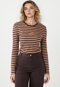 Brązowa i beżowa w paski koszula z długim rękawem, okrągłym dekoltem i haftem konia. Noszona z ciemnoczerwonymi spodniami z wysokim stanem.