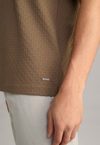 Braun gemustertes kurzärmliges Hemd mit strukturiertem Oberflächenmaterial, das ein kleines silbernes Logo-Tag am Saum trägt. Legere Passform, kombiniert mit hellgrauen Hosen.
