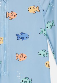 Lys blå rash guard med små, fargerike fiske mønstre i oransje, gult, grønt og blått; inkluderer en glidelås foran for lukking.