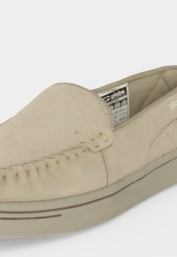 Mocassin beige en daim à enfiler avec coutures mocassin, semelle surélevée et étiquette intérieure visible indiquant la marque et la taille.