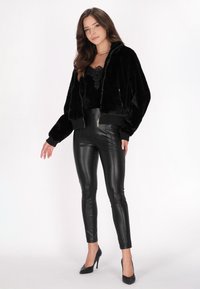 Černá bunda bomber z umělé kožešiny se zipem, žebrovanými cuffs a lemem; kombinováno s lesklými černými přiléhavými legínami a černými vysokými podpatky.