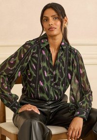 Caurspīdīga melna blūze ar zaļu un purpura dzīvnieku rakstu, apdarināta ar sudraba kniedēm pa priekšu, ar apkakles izgriezumu un puffa piedurknēm.