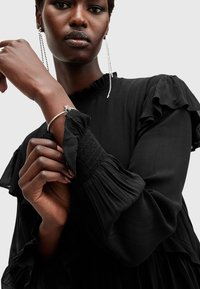 Zwarte, gerimpelde lange jurk met smockmanchetten. Model draagt een zilveren armband en oorbellen, waardoor de textuur van de stof en de ontwerpdetaillering goed tot hun recht komen.