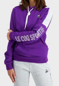 Felpa viola con coulisse bianca e accenti grigi sulle maniche, con la scritta "LE COQ SPORTIF" stampata lungo la manica sinistra. Vestibilità standard.