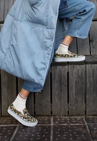 Scarpe slip-on con un motivo a macchie di leopardo nei colori marrone, verde e nero, suola in gomma bianca, indossate con calzini leggeri e jeans blu risvoltati.