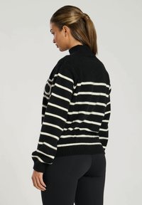 Gestreifter Pullover in Schwarz und Weiß mit hohem Kragen und schmalen Bündchen. Stricktextur mit lockerer Passform, ausgestattet mit einem kreisförmigen Detail an der Seite.