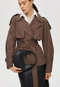 Bruine trenchcoat met grote revers en knoopmanchetten, gecombineerd met een zwarte halve maan handtas, over een witte blouse met kraag en een rok.