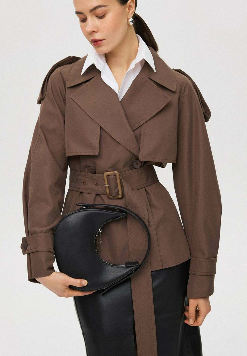 Bruine trenchcoat met grote revers en knoopmanchetten, gecombineerd met een zwarte halve maan handtas, over een witte blouse met kraag en een rok.