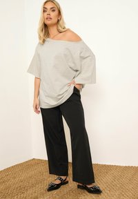 Camiseta de gran tamaño en gris claro con diseño de un hombro al descubierto, combinada con pantalones anchos negros y zapatos planos negros con detalles de hebilla.