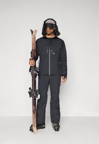 Man wearing black ski jacket, pants, boots, goggles, and gray beanie holding skis standing against a plain white background.