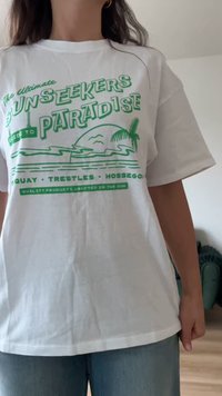 Άτομο που φοράει λευκό t-shirt με πράσινο κείμενο και εικονογράφηση παραλίας που γράφει «Ο απόλυτος οδηγός για τους λάτρεις του ήλιου στον παράδεισο» και ονόματα τοποθεσιών.