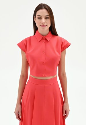 Femme aux cheveux bruns et raides portant un crop top rouge à manches courtes boutonné et une jupe plissée taille haute assortie sur fond blanc.