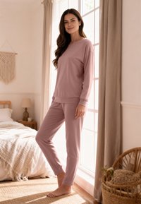 Világos rózsaszín szabadidőruha szett puha anyagból, hosszú ujjú, kerek nyakú felsővel és hozzá illő szűk szárú nadrággal, mezítláb viselve.