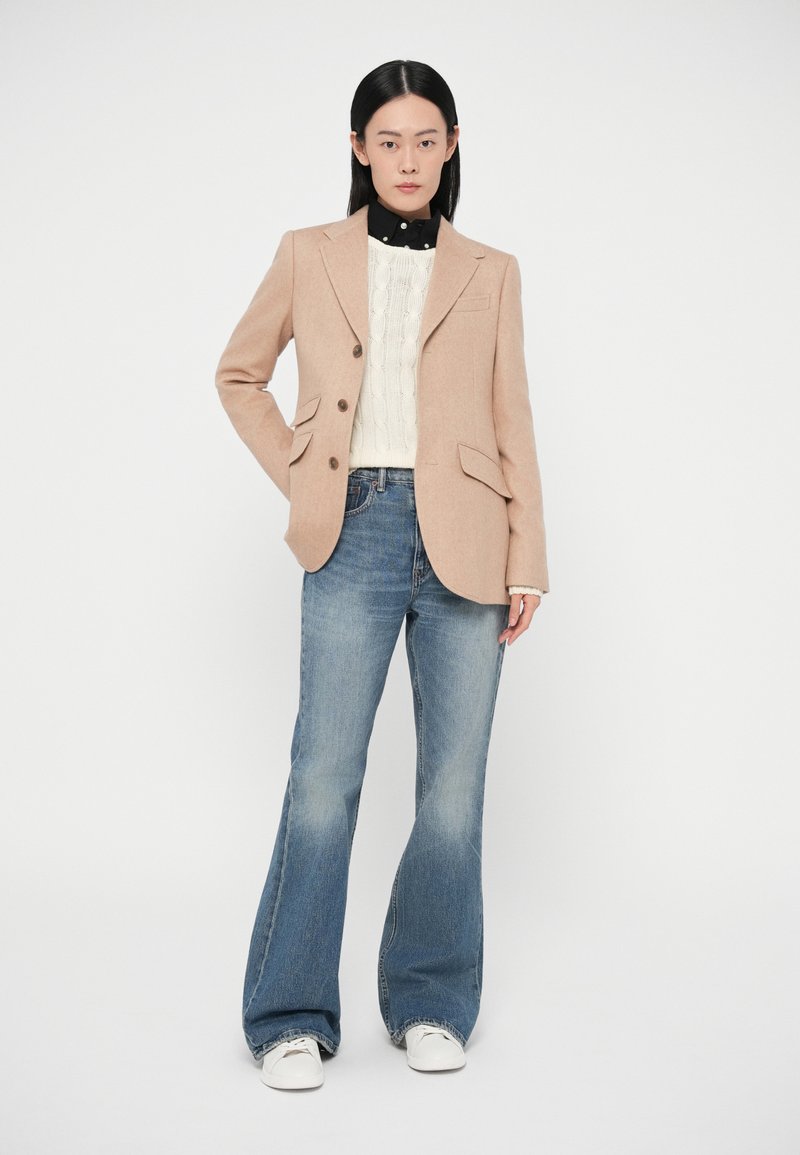Blazer beige con due bottoni e due tasche frontali, indossato sopra un maglione bianco lavorato a maglia, abbinato a jeans svasati blu e sneakers bianche.