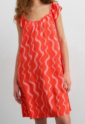 Femme portant une robe rouge sans manches avec des rayures ondulées verticales roses, debout devant un fond clair uni.