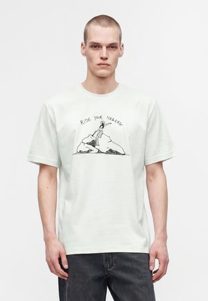 Ung mand med kort hår iført en hvid T-shirt, der viser en tegning af en kvinde på et isbjerg med teksten "Ride Your Iceberg."