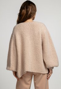 Jasnoróżowy, oversize'owy sweter o puszystej fakturze, z szerokimi rękawami i lekko zaokrąglonym dołem, zestawiony z luźnymi beżowymi spodniami.