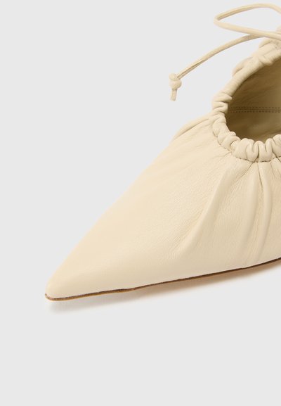 Zapato plano de piel crema con punta afilada y abertura elástica fruncida. Presenta una textura suave y un sutil detalle de costura.
