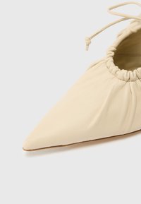 Zapato de cuero crema con punta puntiaguda, apertura elástica fruncida y textura suave. Presenta un acabado liso y detalles mínimos de costura.