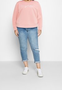 Felpa rosa chiaro con "Tommy Hilfiger" ricamato in bianco, abbinata a jeans blu con risvolto usurato e sneakers bianche.