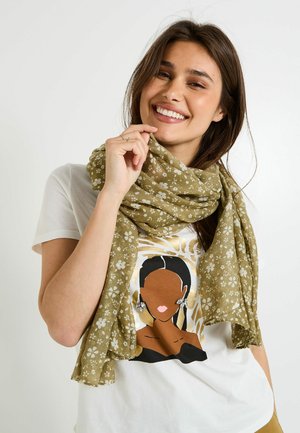 Écharpe vert olive avec des motifs floraux blancs, fabriquée en tissu léger. Portée drapée autour du cou, elle met en valeur une tenue décontractée.
