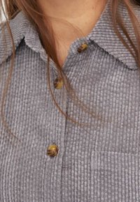 Grå manchester-skjorta med krage, två bruna knappar och synlig ribbad struktur. Inkluderar en bröstfickdetalj.