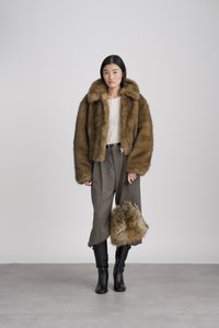 Γυναίκα με καφέ γούνινο μπουφάν και παντελόνι, κρατώντας μια ταιριαστή γούνινη τσάντα, στέκεται μπροστά σε απλό ανοιχτό γκρι φόντο.