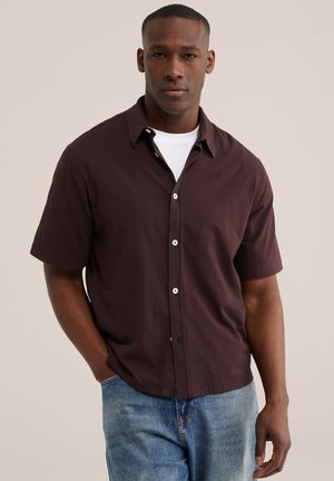 Homme debout, mains dans les poches, portant une chemise à manches courtes marron foncé boutonnée sur un t-shirt blanc et un jean bleu.