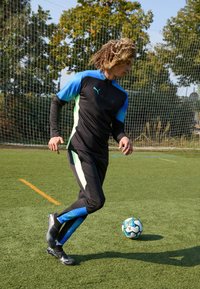 Schwarzes Sportoutfit mit blauen Akzenten, langen Ärmeln und einem Reißverschluss; Spieler tritt einen Fußball auf grünem Kunstrasen.